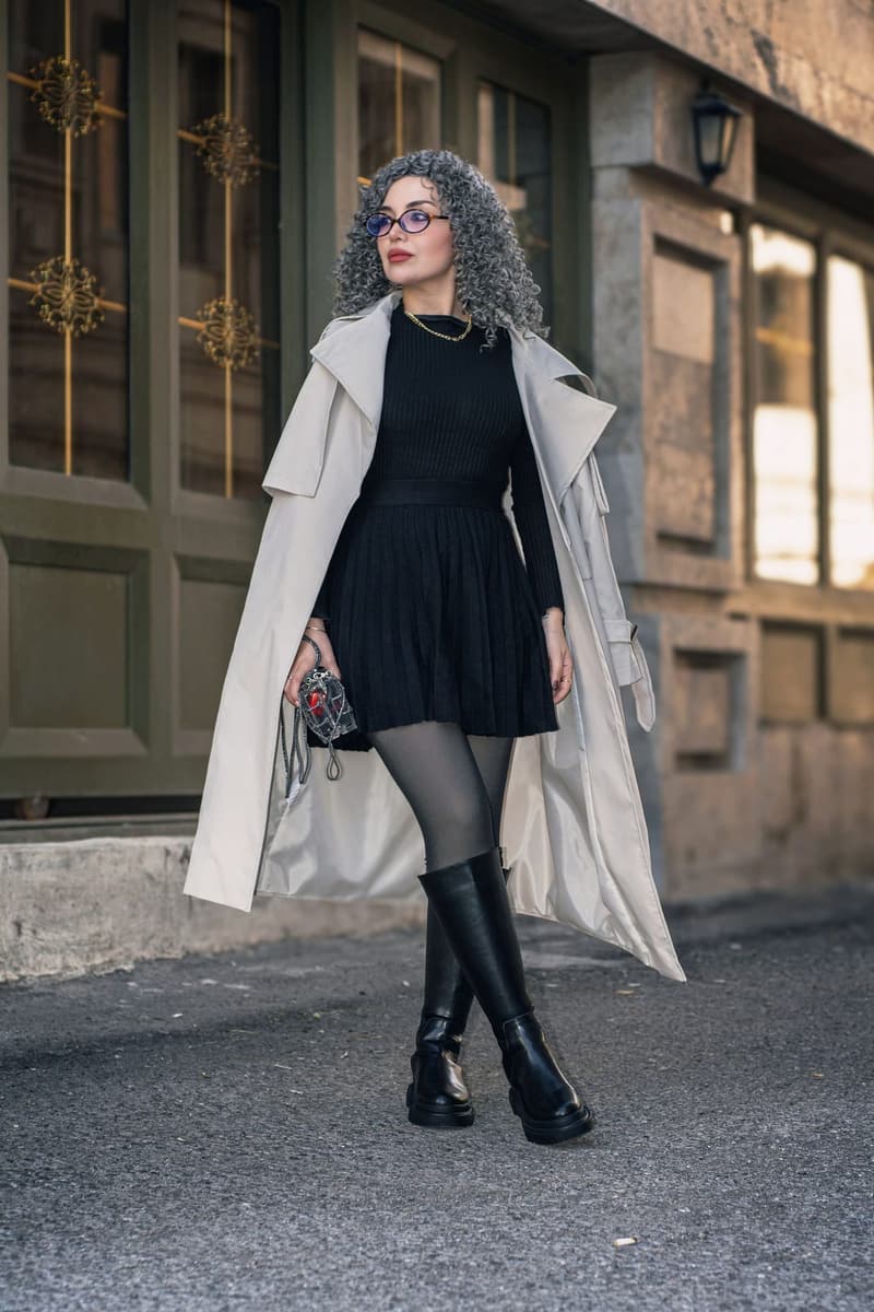 Woman in a long cream trench coat over a black dress and boots, walking past a stone doorway