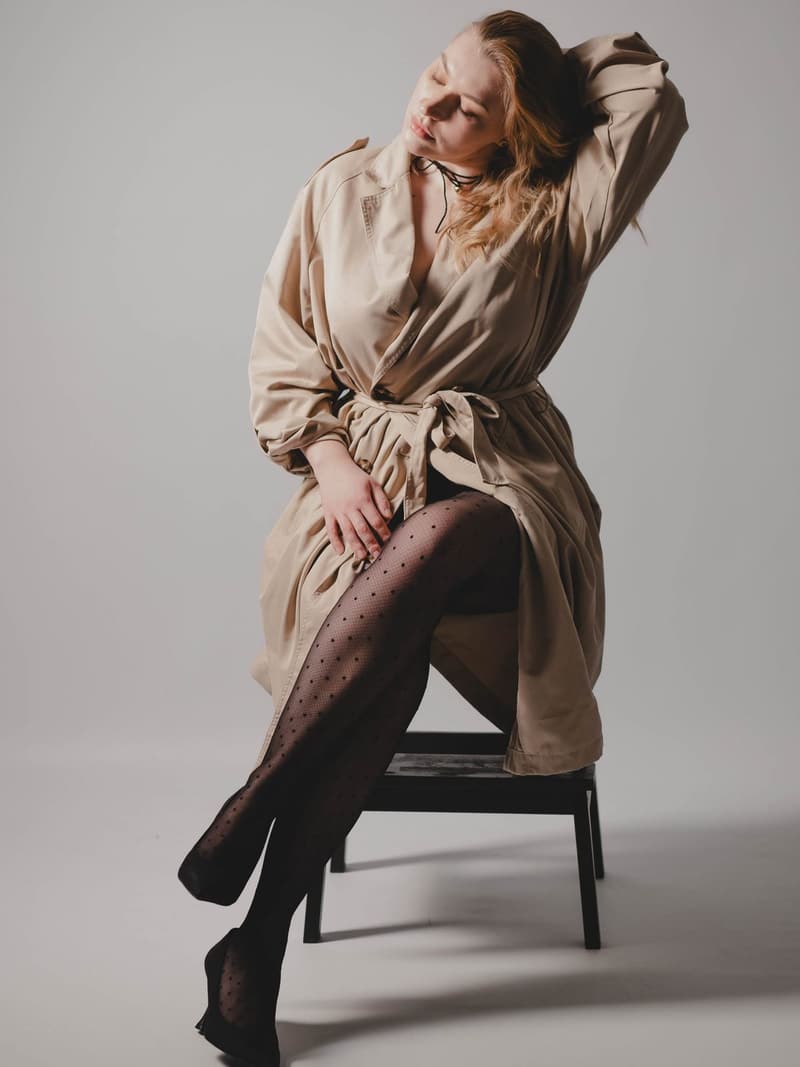 Woman in a beige trench coat seated on a chair in a grey studio, one arm raised